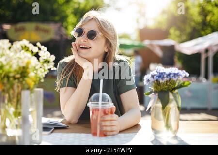 Portrait of young adorable woman drinking lemonade in park surronded by flowes, wearing glasses dreaming of her future career as a beaty blogger. Stock Photo