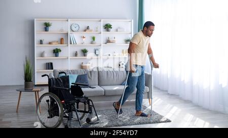 Rehabilitation concept. Young black guy walking with crutches after using wheelchair at home, panorama Stock Photo