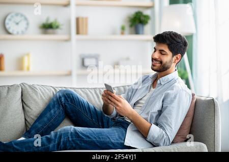 Mobile communication concept. Happy arab man using cellphone, browsing internet, sitting on couch at home, copy space Stock Photo
