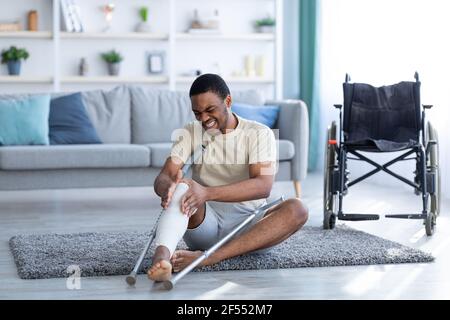 Young black man suffering from pain in plastered broken leg, sitting on floor after falling with crutches, indoors Stock Photo