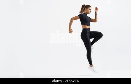 Determined healthy woman doing workout on white background. Fitness trained female raising leg and jumping. Muscular runner exercising and warm up for Stock Photo