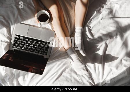 Top view of woman legs in cozy socks, laptop on bed. Girl resting in her bedroom, watching movie online and drinking coffee Stock Photo