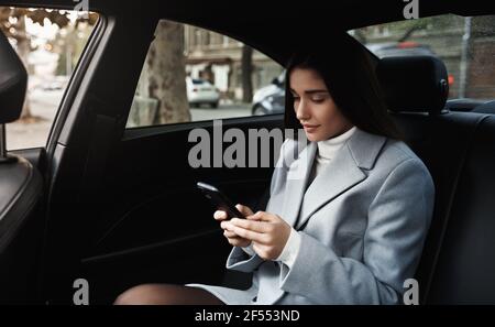 Businesswoman travelling by car on backseat, reading text message on smartphone while driving on meeting Stock Photo