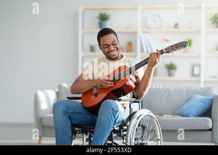 Handicapped black guy in wheelchair playing guitar and smiling at living room. Stay home activities Stock Photo