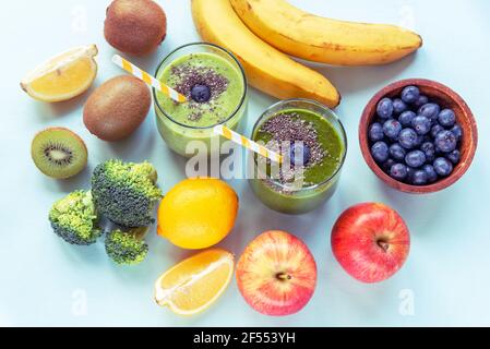 Two different green smoothies in glasses with straws, banana, apples, broccoli, kiwi, lemon and blueberries in a bowl. Healthy ingredients, bright blu Stock Photo