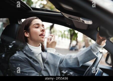 Businesswoman sitting in car and using lipstick, looking at herself in rear-view mirror to check on makeup Stock Photo