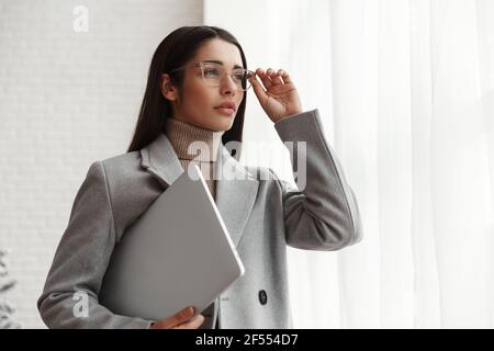 Portrait of a confident businesswoman standing inside office building with laptop computer. Female entrepreneur in glasses looking outside window Stock Photo