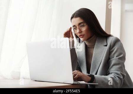 Young businesswoman in glasses sitting in office building, working on laptop. Female entrepreneur looking busy and serious, reading something on Stock Photo