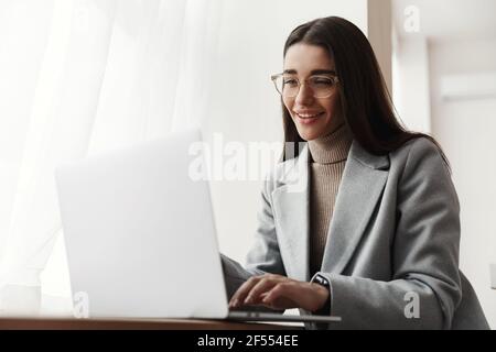 Businesswoman sitting in grey coat and glasses and working on computer. Young woman using laptop and smiling, freelancing at co-working space Stock Photo