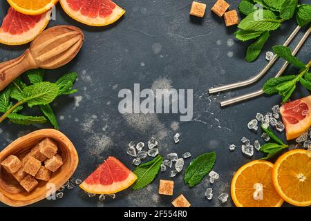 Grapefruit and orange slices, mint, cane sugar, ice, cocktail tubes, juicer or squeezer on black stone old background. Ingredients for making summer b Stock Photo