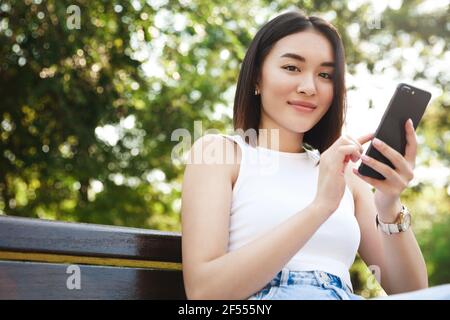 Stylish asian girl sitting in park and using smartphone, smiling at camera. Beautiful modern woman with cell phone resting on bench outdoors Stock Photo