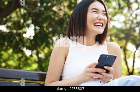 Happy asian girl sitting on bench and using mobile phone. Girl resting in park and browsing social media on smartphone, laughing at someone Stock Photo