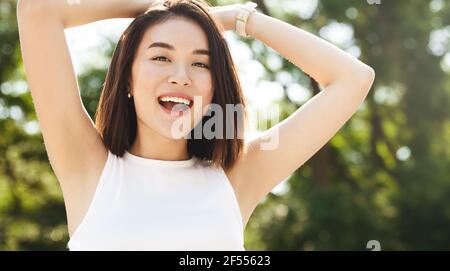Close-up of happy asian woman posing in park, smiling and looking at camera, holding hands behind head relaxed. Girl enjoying sunny day outdoors Stock Photo