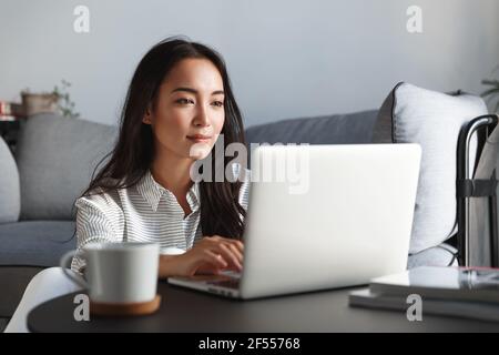 Young ambitious asian girl working remote from home, looking at laptop screen and smiling. Woman checking mail or researching while telecommuting Stock Photo