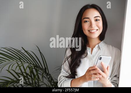 Portrait of happy attractive asian girl smiling at camera, standing near window with smartphone. Woman spending time at home, waiting for phone call Stock Photo