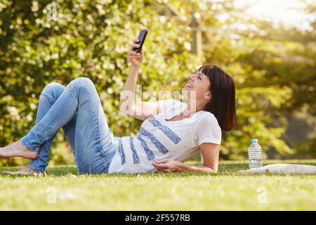 Full body portrait of happy woman lying in grass taking selfie with smart phone Stock Photo