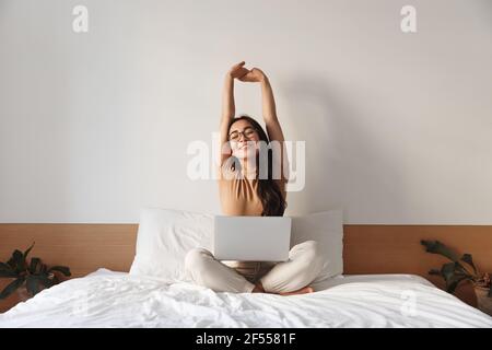 Portrait of satisfied asian girl sitting on her bed with laptop and stretching hands up satisfied, finish work project and looking delighted. Female Stock Photo