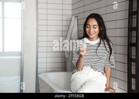 Photo of young asian woman sitting in bathroom with smartphone. Girl using phone and sit on bathtub, smiling and reading message Stock Photo