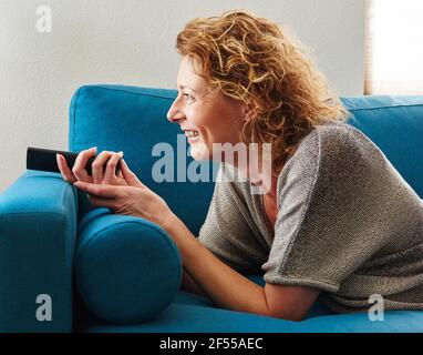 Close up side portrait of smiling woman lying down on couch with remote control Stock Photo