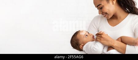 African Mother Feeding Baby Standing On White Background, Panorama, Cropped Stock Photo