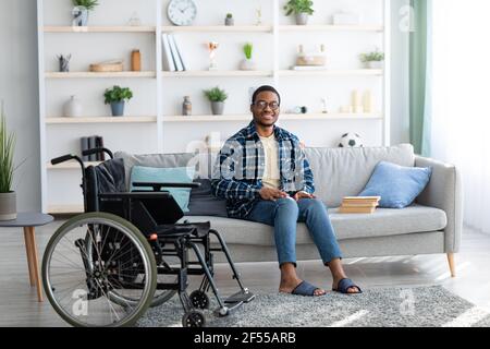 Happy disabled black guy sitting on sofa with books, empty wheelchair standing nearby, full length portrait Stock Photo