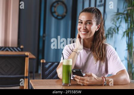 Attractive smiling woman on vacation, enjoying paradise resort, sit in cafe using mobile phone and drinking healthy smoothie, look camera, self-care Stock Photo