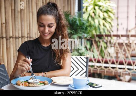 Food and tropical vacation concept. Beautiful tanned woman eating at hotel cafe, sit outdoors on terrace and trying Bali breakfast, drinking morning Stock Photo
