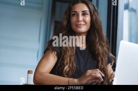 Smiling businesswoman working from home, sitting in room and using laptop, looking away dreamy. Self-employed woman stay indoors and freelancing with Stock Photo