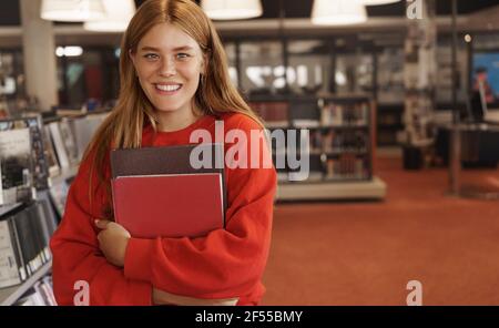 Education, young generation and people concept. Redhead girl studying hard for exams, cramming in library, holding books, standing in bookstore near Stock Photo