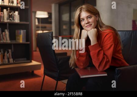 Portrait of young attractive redhead female student, sit armchair lean on arms and smiling, looking camera, discussing novel with bookclub in Stock Photo