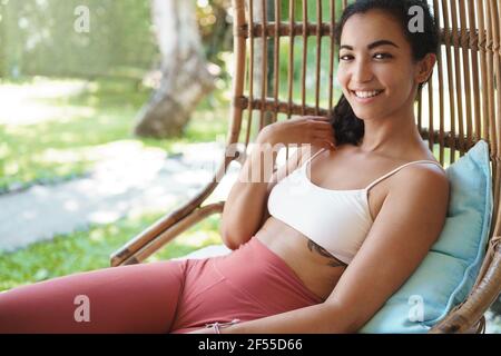 Woman feeling energized and upbeat after morning yoga exercises in garden. Attractive smiling female in sports bra, leggings lying rattan armchair Stock Photo
