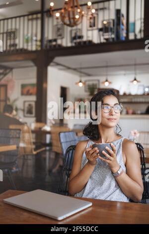 Vertical shot successful attractive woman freelance digital nomad enjoy summer nature urban view sit near closed laptop cafe table hold coffee mug Stock Photo