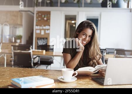 Charming happy smiling alluring tanned woman in cafe studying prepare homework reading interesting book lean table grinning camera near opened laptop Stock Photo