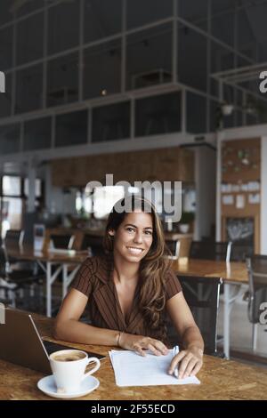 Vertical shot successful charming young brazilian businesswoman, entrepreneur sit in cafe, co-working urban space with opened laptop, drink cappuccino Stock Photo