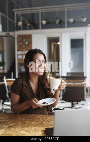 Vertical shot gorgeous hispanic woman taking coffee break, sit alone co-working space or cafe, laptop opened on table, she holds cup as tastes Stock Photo