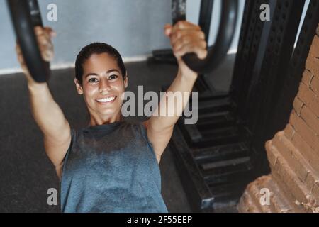 Upper-angle shot attractive sweaty sportswoman using gym equipment to gain perfect abs, good body shape, push-up on rings, pumping biceps, smiling as Stock Photo