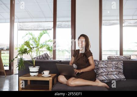 Co-working, business and finance concept. Attractive happy young hispanic woman in fancy dress, lying on sofa near plants and terrace, holding laptop Stock Photo