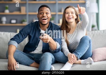 Cheerful multiracial couple having fun at home, watching comedy movie on tv Stock Photo