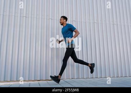 Male athlete with mobile phone running on sidewalk by wall during training Stock Photo