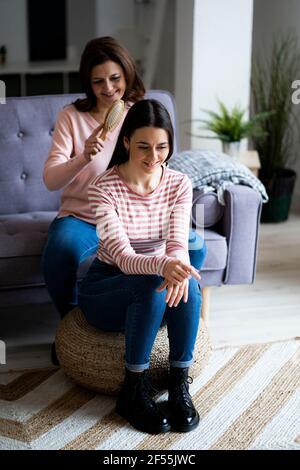 Smiling mother combing daughter's hair at sofa in living room Stock Photo