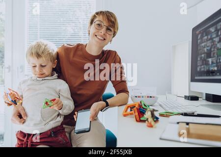 Smiling mother with son sitting on lap playing with toys in home office Stock Photo