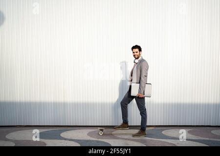 Smiling male business professional with laptop standing on longboard by wall Stock Photo