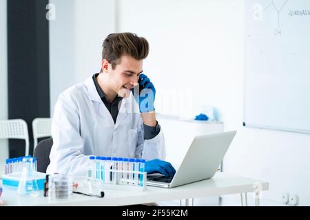 Male scientist talking on smart phone while using laptop at desk in laboratory Stock Photo