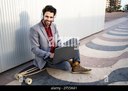 Smiling businessman with laptop sitting on longboard in front of wall Stock Photo