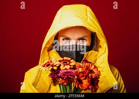 Mid adult woman wearing hood clothing and protective face mask staring while standing with bunch of flowers against red background Stock Photo