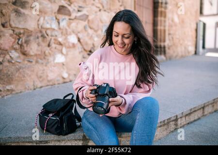 Beautiful woman smiling and holding camera while sitting on staircase against wall Stock Photo