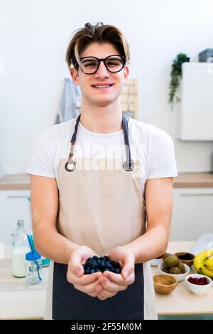 Smiling young man in apron holding fresh blueberries while standing in kitchen Stock Photo