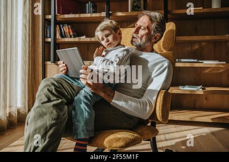 Boy E-learning through digital tablet while sitting on father's lap on chair at home Stock Photo