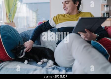 Smiling man with digital tablet stroking cat while sitting on sofa in living room Stock Photo