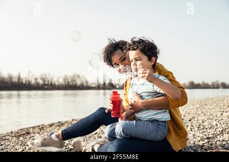 Boy playing with bubble wand while sitting on mother's lap during sunny day Stock Photo